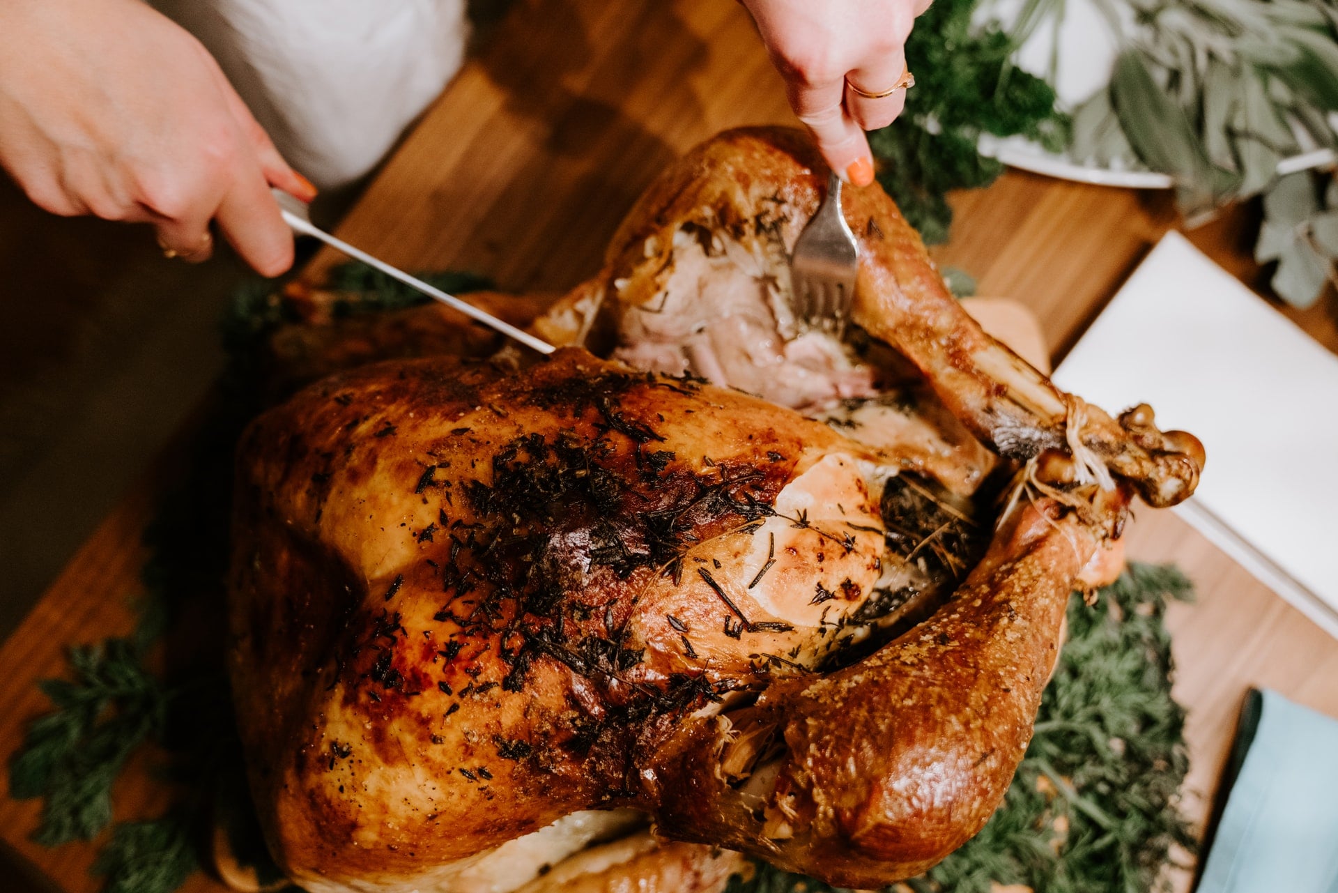 Woman's hands cutting into a golden brown roasted turkey covered in herbs and set on a wooden oak table